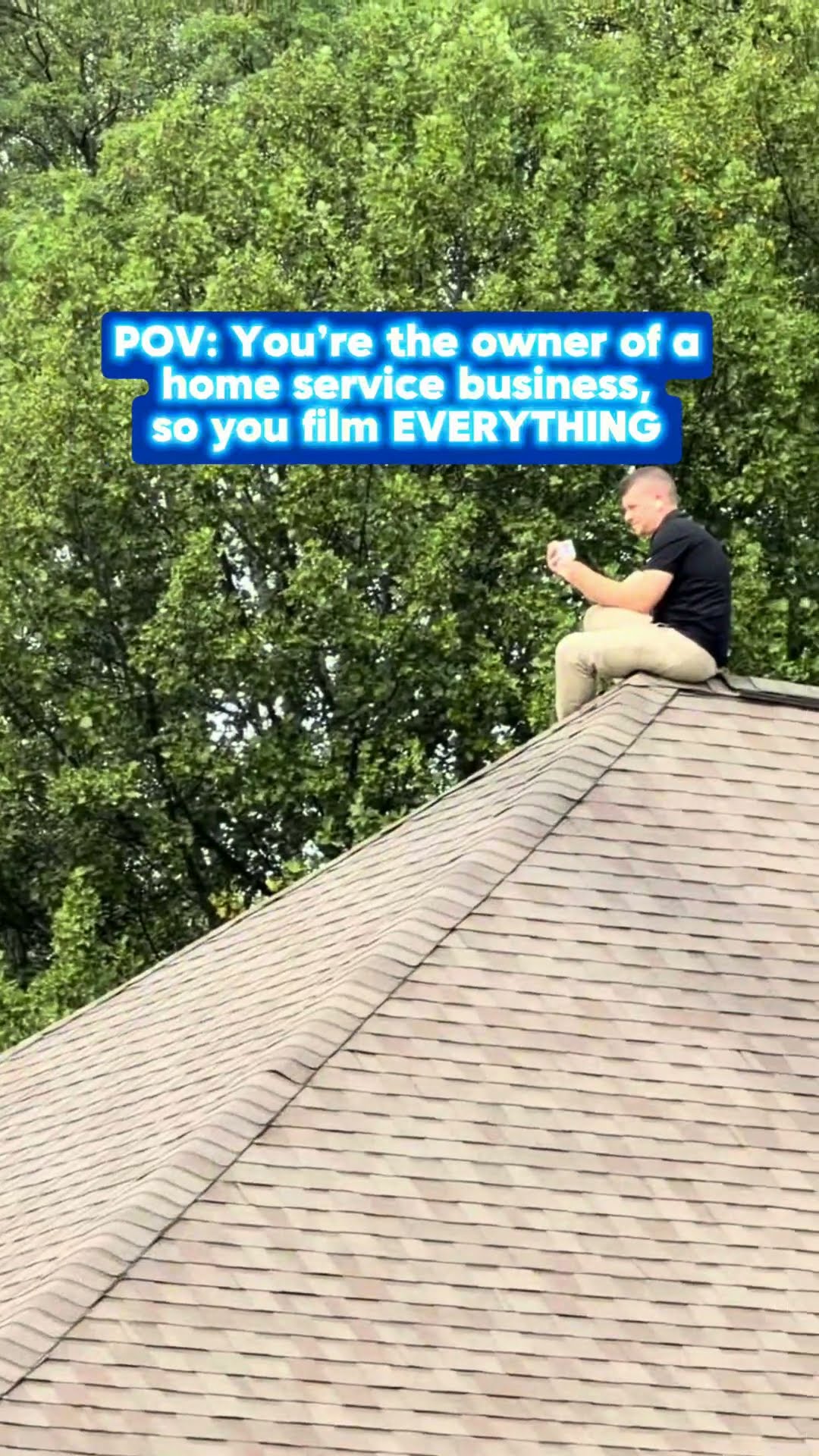 A man in a black shirt sits on the peak of a shingled roof filming on his phone while a red crane truck sets up in a parking lot below for an equipment lift.