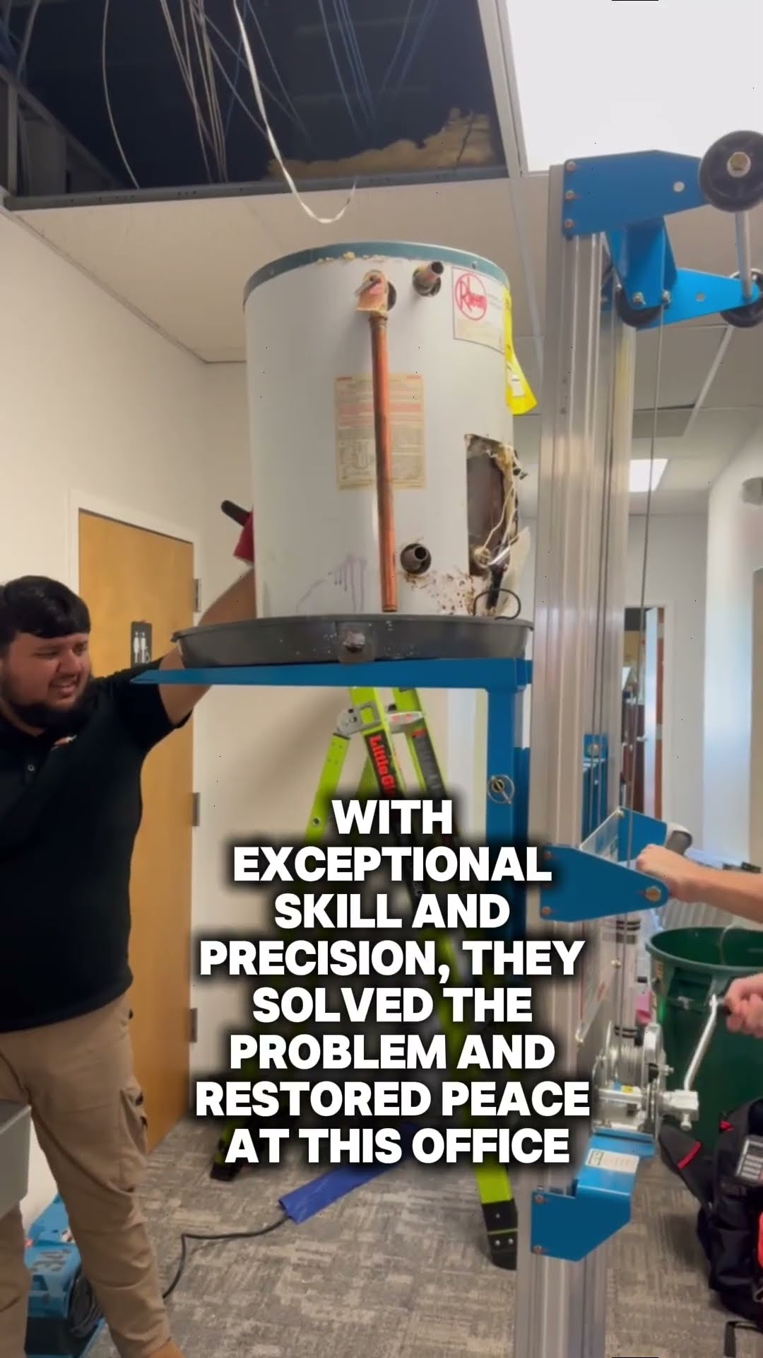 A commercial office water heater job: a Rheem tank water heater is raised on a blue material lift into an open drop ceiling while a tech operates it, with wet-vac and tools on office carpet.