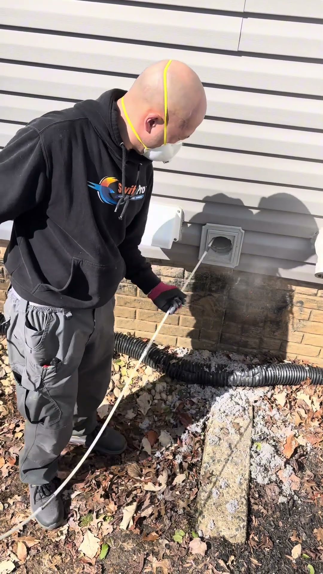 Dryer vent cleaning outside a house: a tech in a black SwiftPro hoodie and dust mask runs a rotary brush rod into an exterior dryer vent on vinyl siding over brick, with lint blowing out onto the mulc