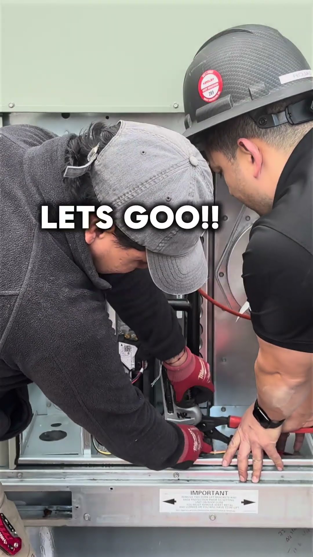 Two techs work inside the gas section of a commercial rooftop package unit, one in a gray cap and hoodie and one in a black hard hat.