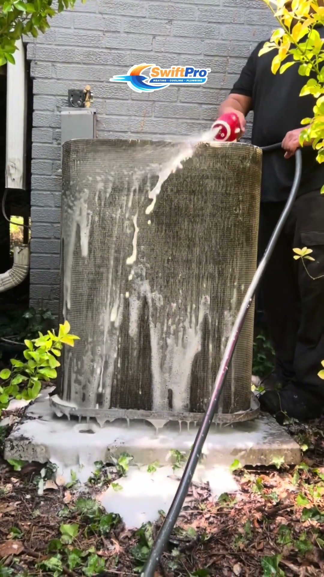 Outdoor AC condenser cleaning: leaves and mulch packed inside the base, then a tech in black with red gloves sprays foaming coil cleaner and rinses the coil with a hose beside a brick house.