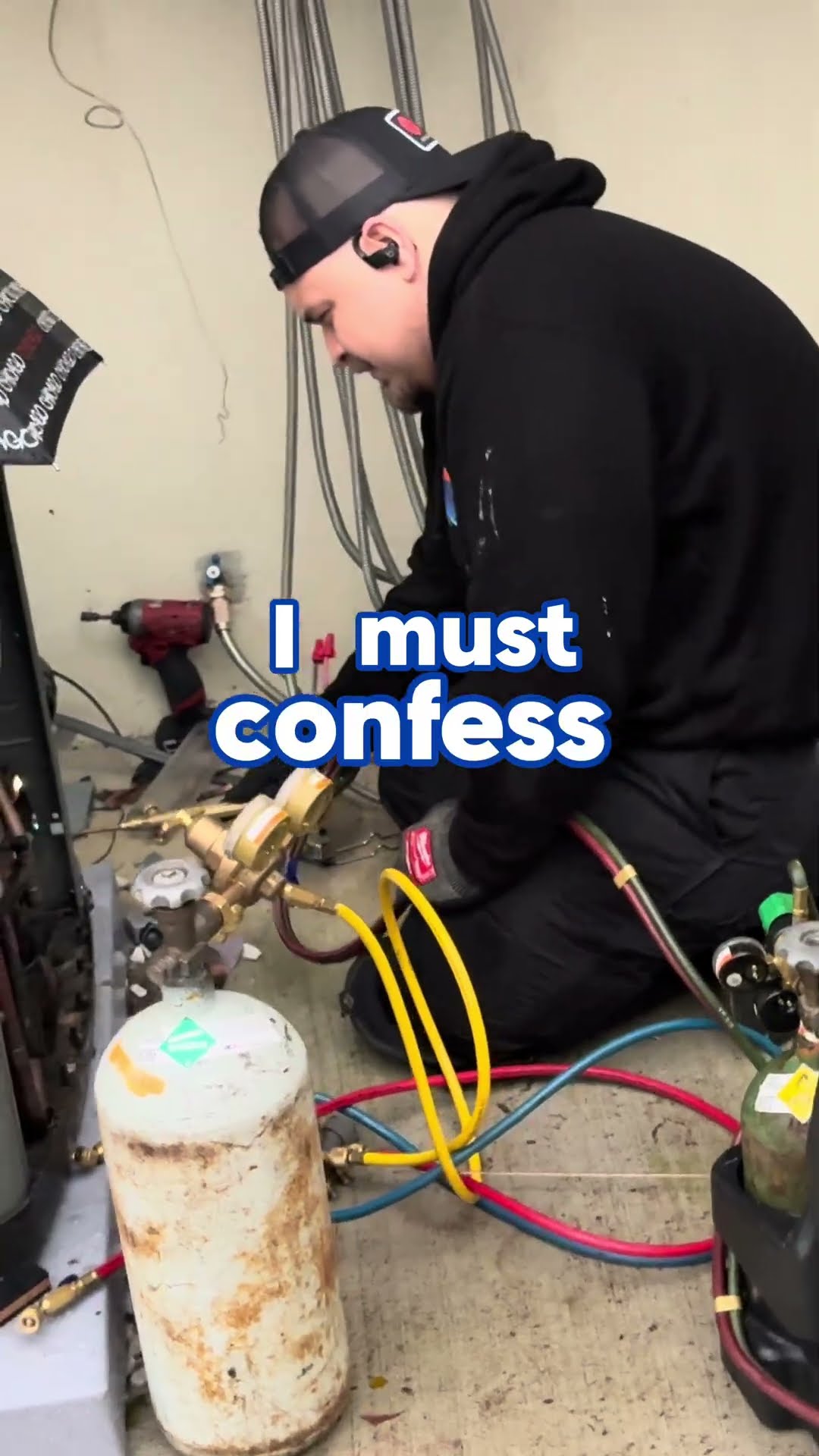 A tech in a backward black trucker cap and hoodie kneels at an AC condenser with nitrogen and torch tanks and a gauge manifold, set up to braze.