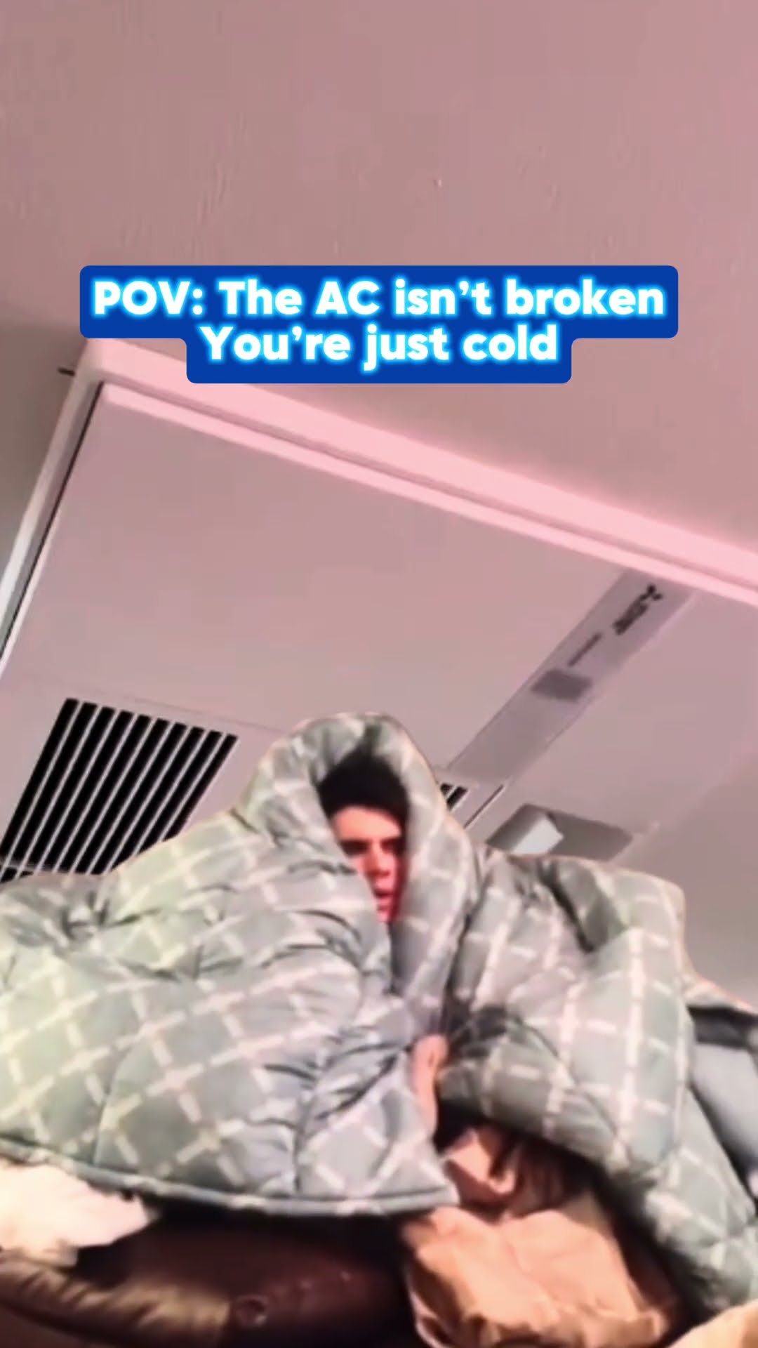 A person bundled in a blanket on a couch under a ceiling air vent, with the caption the AC isn't broken, you're just cold.