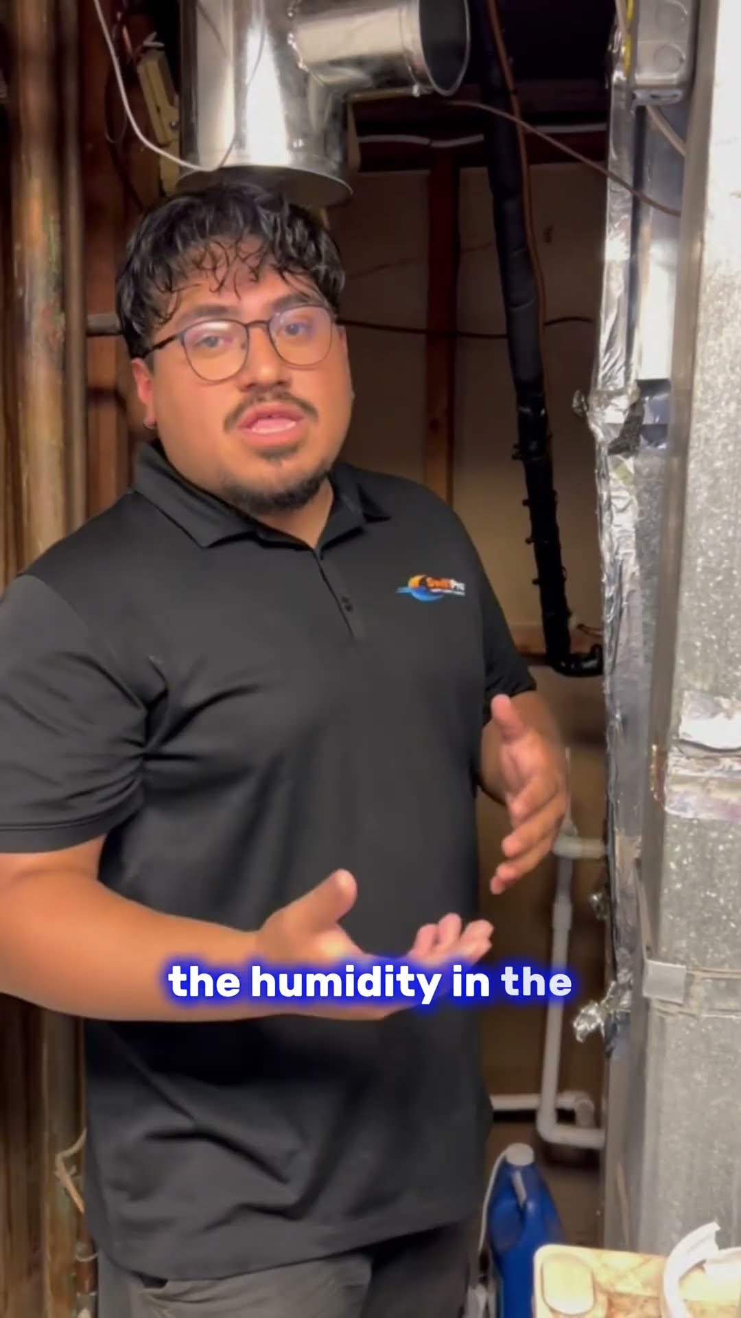 The same tech in a black SwiftPro polo stands in an unfinished basement by the air handler and ductwork, explaining how a clogged AC condensate drain and humidity can stop the system, holding a drain