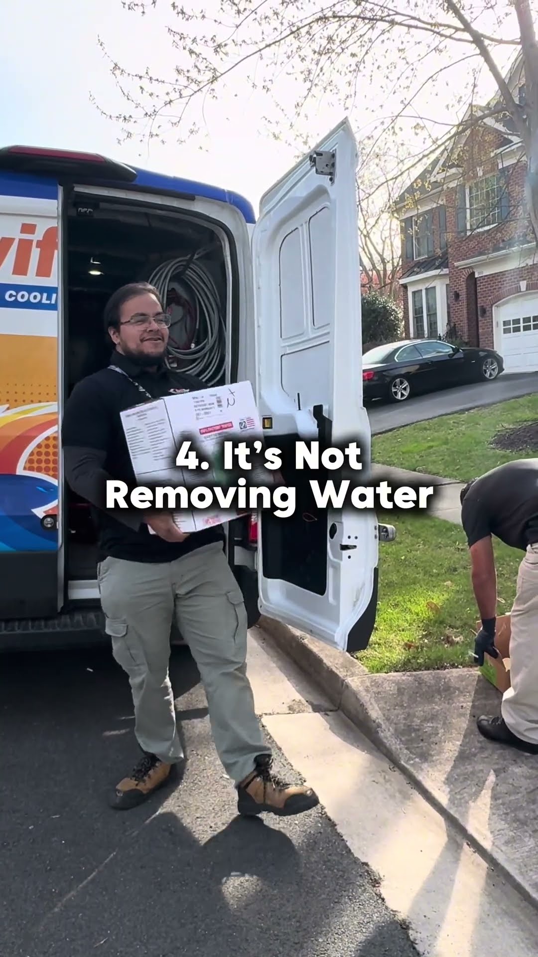 A tech in a black SwiftPro shirt carries a boxed sump pump from a wrapped SwiftPro van in a residential driveway.