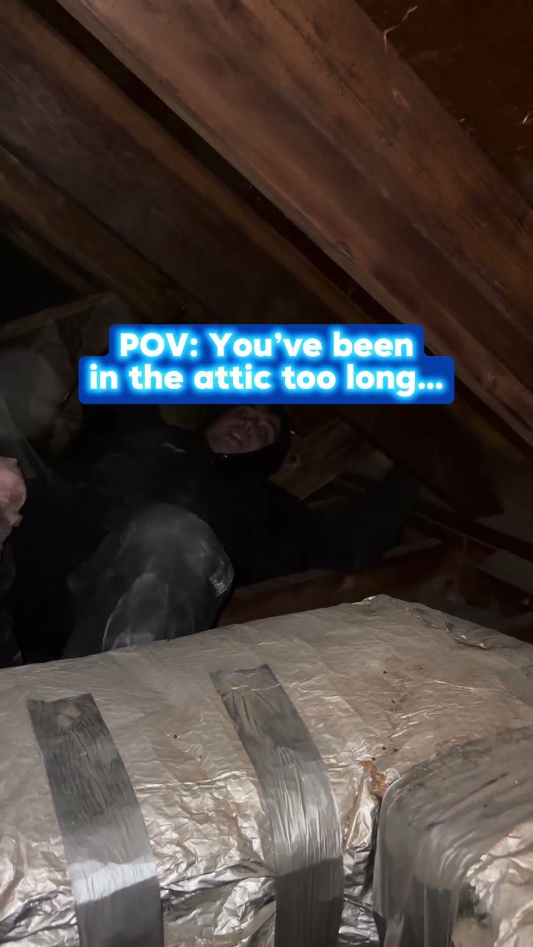 A dim attic with foil-wrapped flex duct in the foreground and a person in dark clothing and a face covering crouched among the rafters.