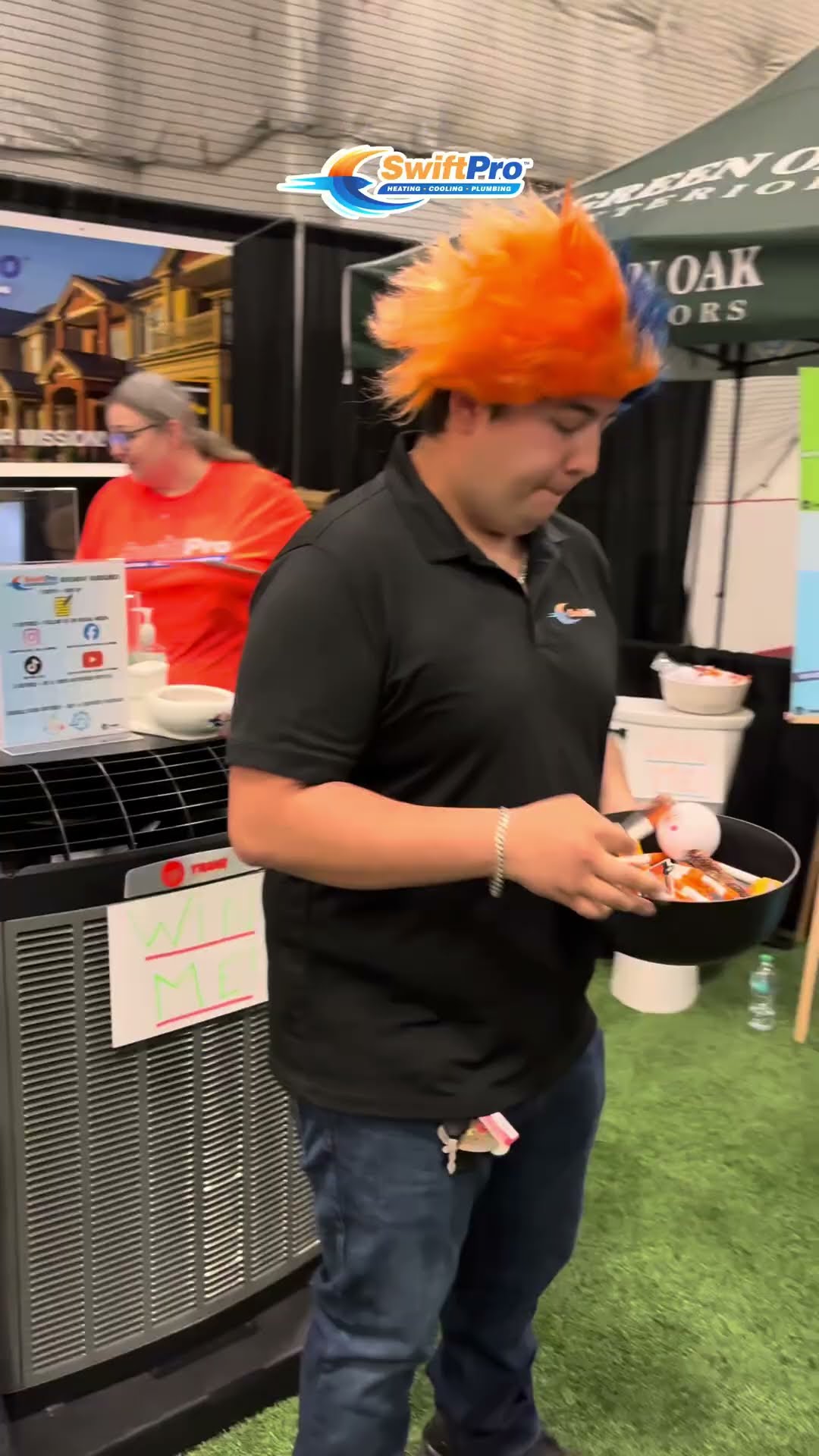 Home show recap at an indoor expo hall on turf: a SwiftPro staffer in a black polo and a spiky orange wig carries a bowl of giveaways at the SwiftPro booth beside a Trane condenser display, with other