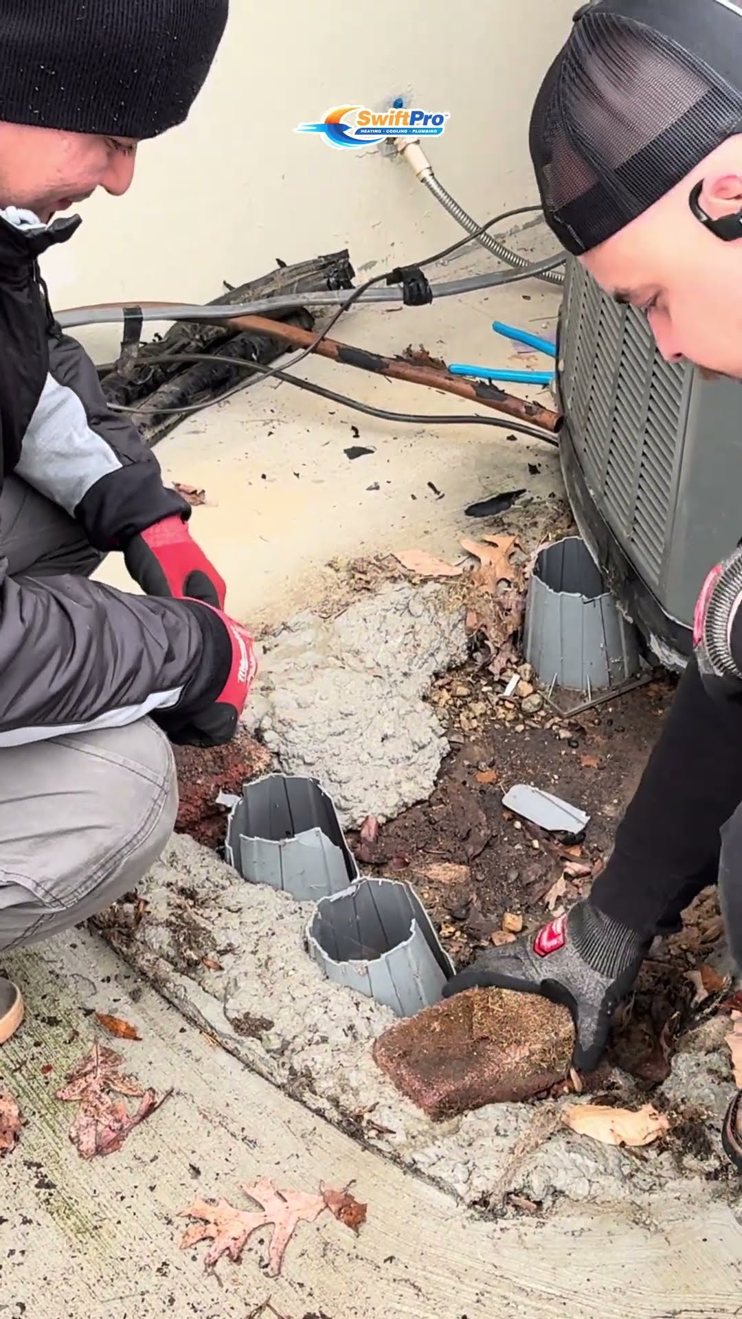 Two techs set a new AC condenser on a concrete patio beside a house in winter, with a refrigerant recovery machine and hoses nearby.