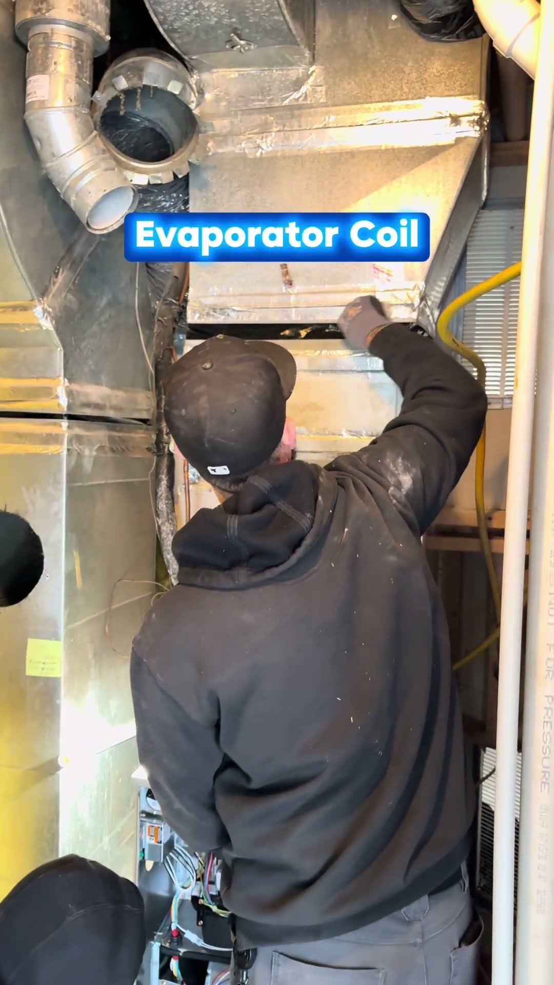 A tech in a black cap and hoodie reaches into the evaporator coil cabinet above a furnace in a mechanical room, labeled on screen.