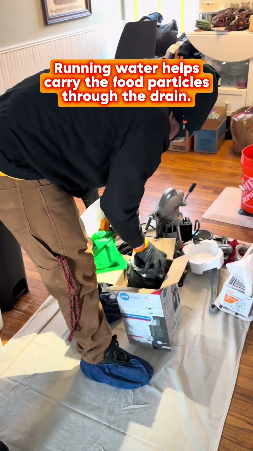 Garbage disposal replacement under a kitchen sink: a tech in a black hoodie removes the old disposal into a blue parts bin on drop cloths, with the new disposal box and tools on the floor.