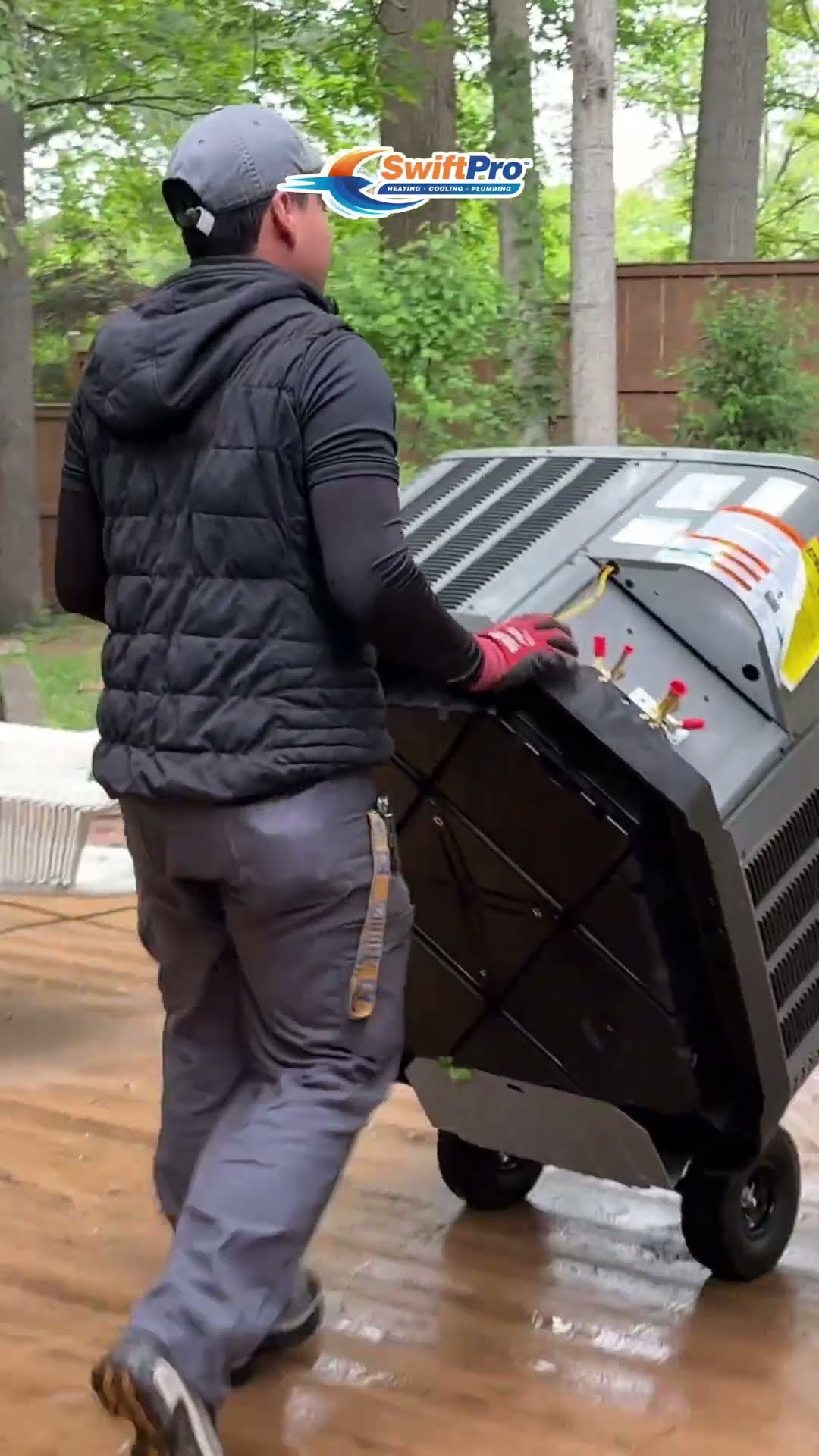 AC replacement: a tech wheels a new condenser on a hand truck across a wet deck, another tech reads digital manifold gauges on the old condenser by a brick wall, and the new condenser sits finished on