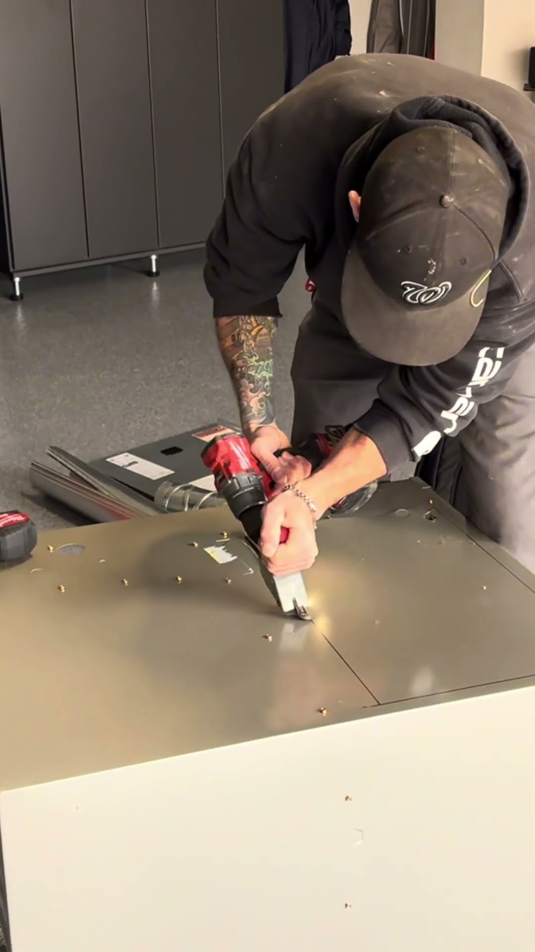 A tattooed tech in a black cap and hoodie cuts an opening in the sheet metal top of an HVAC cabinet with a drill-powered metal shear in a garage.